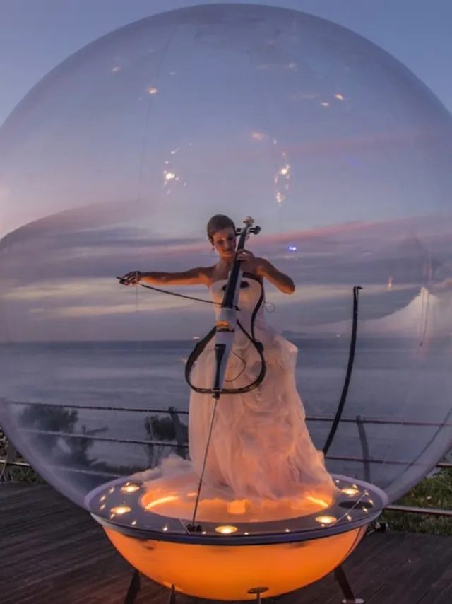 Cellist in a white gown playing inside a glowing bubble above the sea