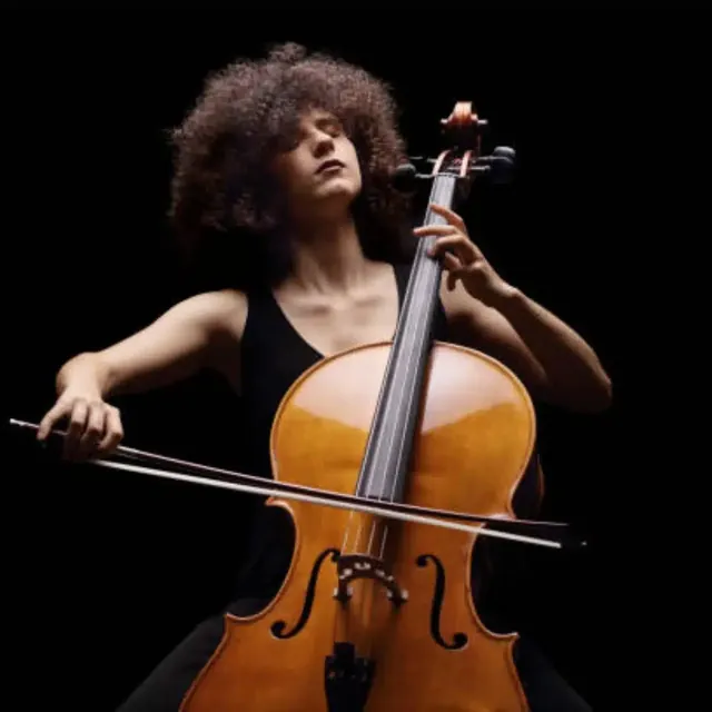 Cellist with curly hair playing a cello on a dark stage